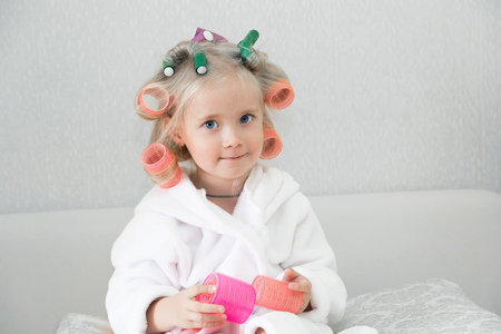 Girl with curlers hairpins in her hair. A little girl in a white coat spun pink curlers into her hair.の写真素材