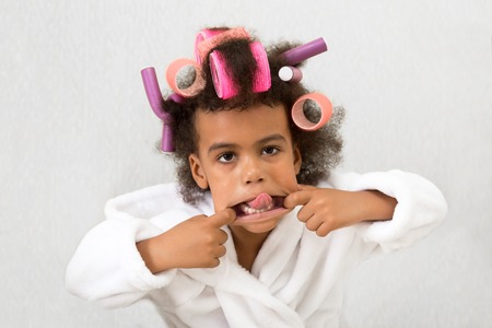 Black girl with curlers with hairpins in her hair. A little girl in a white coat spun pink curlers into her hair.の写真素材