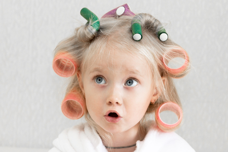 Girl blonde with curlers in her hair. Little girl in white coat with hairpins in her hair on her head.の写真素材