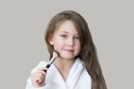 Caucasian girl pains her face with make-up powder. Little girl playing with makeup while sitting on the couch.の写真素材