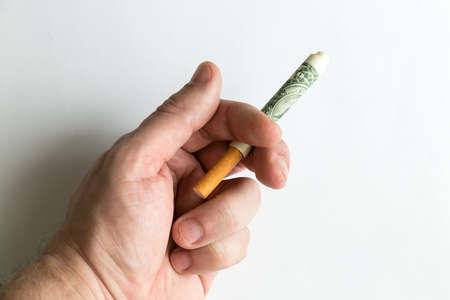 A hand is holding a cigarette rolled into a dollar bill. Smoker's hand holds a cigarette hidden in paper money ...の写真素材