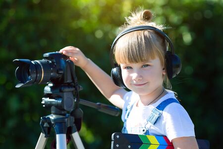 Portrait of a child with a TV clapper. A little girl in headphones with a TV in her hands is standing next to a camera on a tripod.の写真素材
