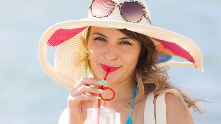 Sexy brunette with juice on the beach. Girl in a hat, blue bikini and a glass juice on vacation.の写真素材