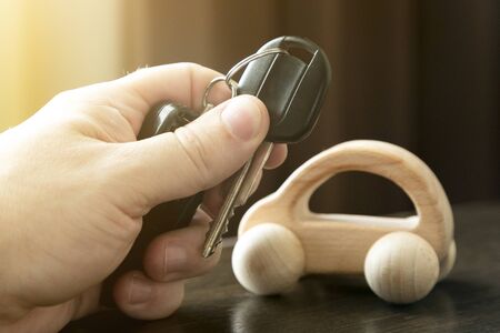 Hand with car keys on a background of the car. Male hand holds car keys next to a small wooden car.