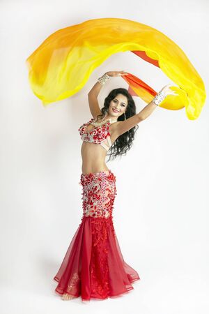 The dancer of east dances with bright scarfs fabrics. A brunette woman performs a belly dance waving in air with colored shawls on a white background.の写真素材