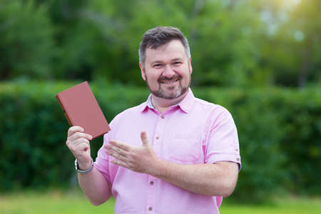 Happy middle-aged Caucasian man in a red shirt with pleasure shows a new book. Satisfied man advertises a new book.の写真素材
