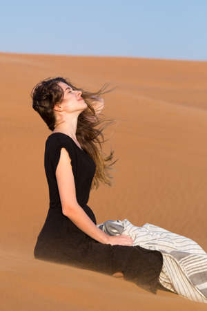 A woman of 20-25 years old in a black dress sits on warm sand of the desert.の写真素材