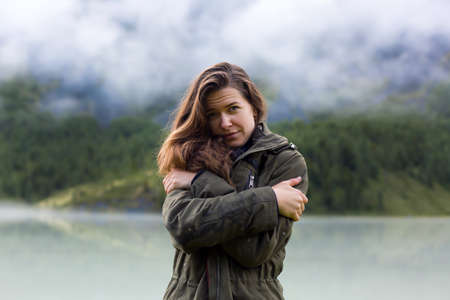 Girl tourist freezes from damp rainy weather in the mountains. A young girl hugs herself by the shoulders from the cold weather in the morning in the mountains.の写真素材