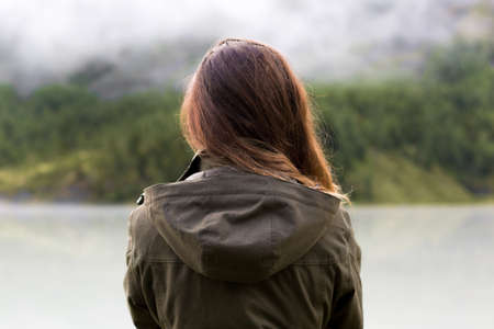 A girl in a jacket looks at the mountains, see from behind. A woman with long hair stands with her back, looks at the mountains, rainy weather.の写真素材