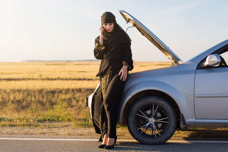 Muslim woman calling help phone near open hood of car.