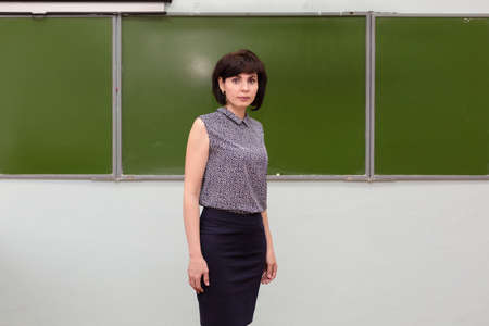 A brunette schoolteacher stands at the blackboard with her arms folded.
