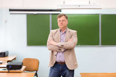 The serious headmaster stands in the classroom at blackboard with his arms folded.の写真素材
