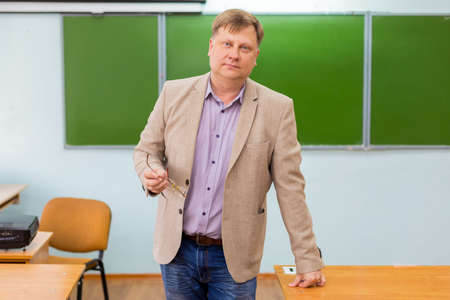 Male teacher blonde 40-45 years old stands with glasses in his hands at the blackboard in the classroom.の写真素材