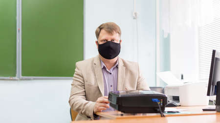 A teacher sits at a desk wearing protective black mask in a school classroom.の写真素材