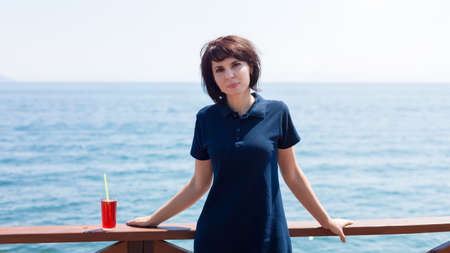 A smiling brunette with a glass of juice is standing on the pier by the sea.の写真素材