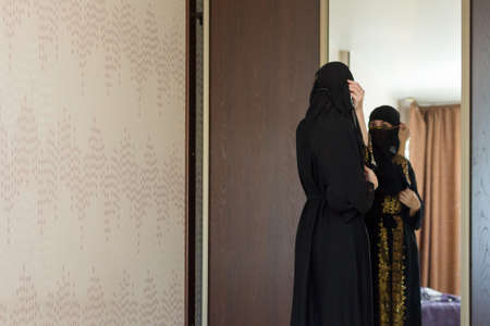 A Muslim woman dresses up in front of mirror and puts on a burqa in her apartment.