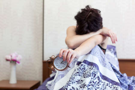 A sleepy woman is sitting on the bed with her face buried in her knees and holding an alarm clock in...