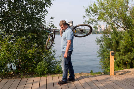 A frustrated man carries a broken bicycle on his shoulders by river in the park.の写真素材