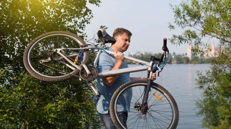 A frustrated man carries a broken bicycle on his shoulders by the river in the park.