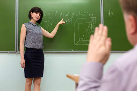 The teacher at the blackboard with a smile asks the answer from the student who pulls his hand.の写真素材