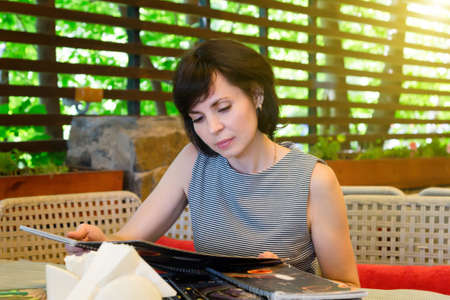 A brunette woman in a restaurant flips through the menu choosing a dish while sitting at a table.