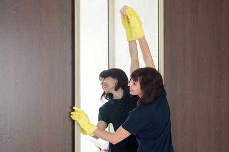 A woman washes the mirror of a high wardrobe in an apartment standing on tiptoe trying to reach...