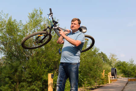 A frustrated man in the park carries a broken bicycle on his shoulder.