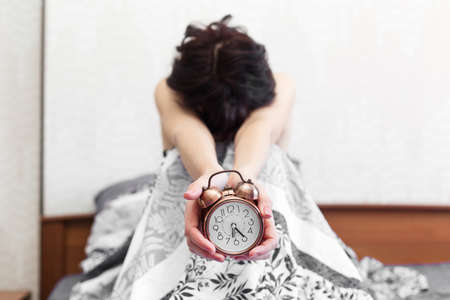sleepy woman is sitting on the bed with an alarm clock in her hands unable get up for work.の写真素材
