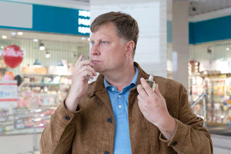 An adult serious man blond in a brown jacket sniffs perfume in a shopping center trying to catch the...