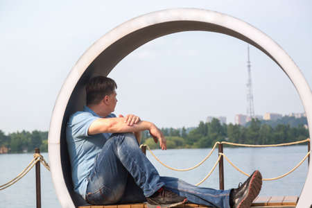 An adult man sits on a creative bench in form of a circle and looks at the calm surface of the...