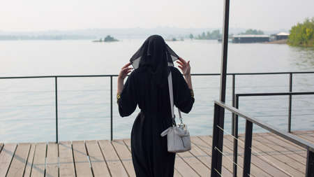 A Muslim woman in national dress walks a wooden bridge to the river, rear view.の写真素材