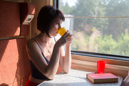 An adult brunette compartment is drinking tea at the table enjoying a train ride.