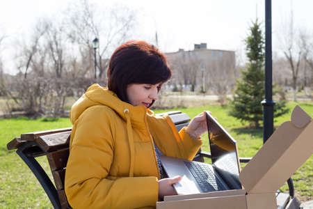 A happy brunette woman in a yellow jacket street eagerly opens a new laptop.の写真素材