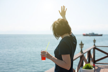 The girl is waving to a ship sailing distance while standing on the shore with a glass of juice.の写真素材