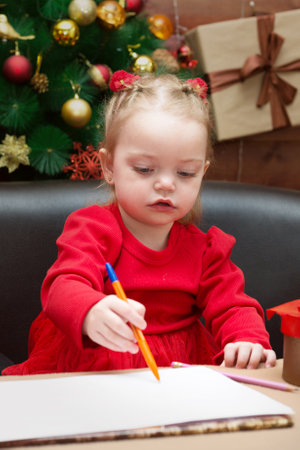 A little girl in a red dress draws on the background of a decorated Christmas tree and presents writes a letter to Santa Claus.の写真素材