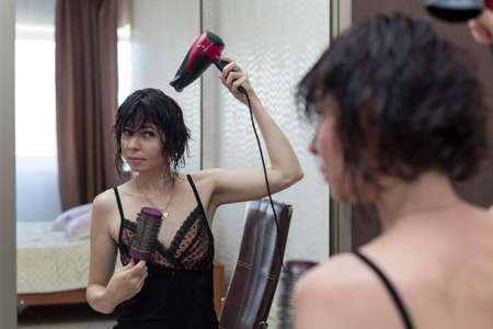 Young woman getting ready for a date, drying her head with a hair dryer in front of a mirror.の写真素材