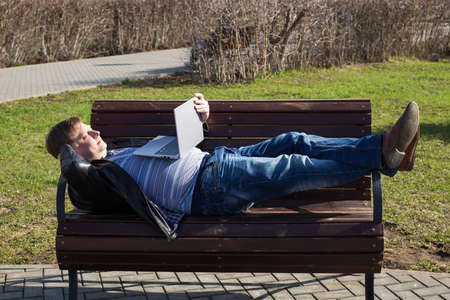 An adult man lay down on a bench in the park and watches a movie on a laptop.の写真素材