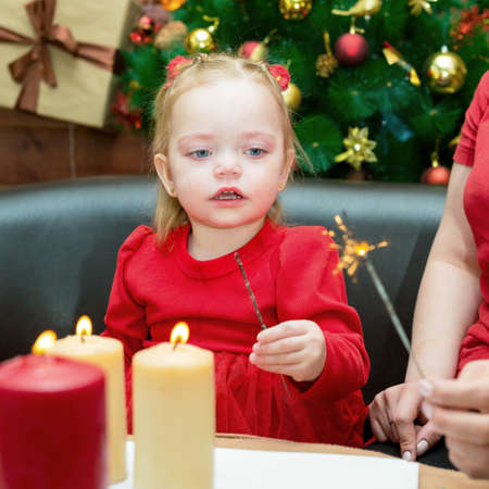 A little girl with her mother lights sparklers in anticipation new year on the background of a green Christmas tree.の写真素材
