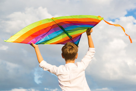 A 9-year-old boy with a bright colorful kite stands high on a mountain against background of beautiful clouds and looks into distance.の写真素材
