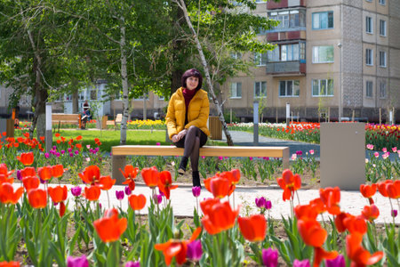 A woman in a bright yellow jacket is sitting on a bench in a city park among tulips.の写真素材