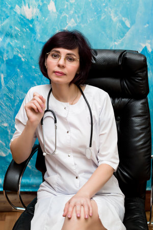 Woman doctor sits in a chair in office against blue wall.の写真素材