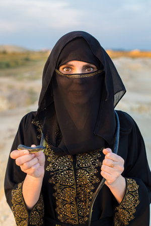 A Muslim woman in national dress holds a snake in her hands while standing desert.