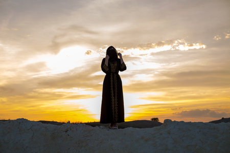 An Islamic woman prays while standing on a mountain sunset. Religious tradition evening prayer.の写真素材