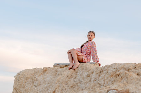 A girl in a light dress sits barefoot on a high hill on a summer evening.の写真素材
