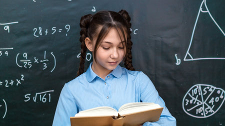 Beautiful teenage schoolgirl reading a textbook for teacher while standing at a blackboard at...
