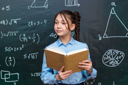 Beautiful teenage schoolgirl reading textbook for a teacher while standing at a blackboard at...