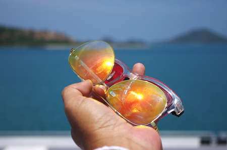 Hand holding colorful sunglasses against and blue sunny sky on the beach with beautiful of reflexの写真素材