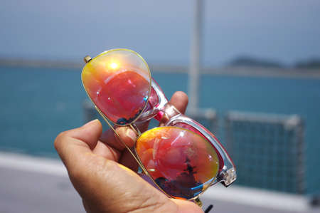 Hand holding colorful sunglasses against and blue sunny sky on the beach with beautiful of reflexの写真素材