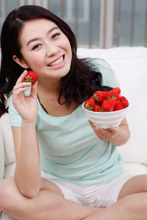 Young woman sitting on a couch, holding a bowl of strawberriesの写真素材