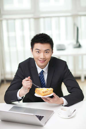 Portrait of businessman sitting at desk by laptop,holdig sandwhichの写真素材